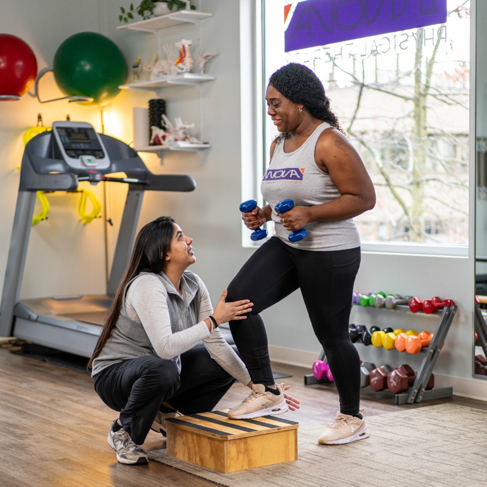 A patient holding hand weights steps up onto a block with assistance from a therapist