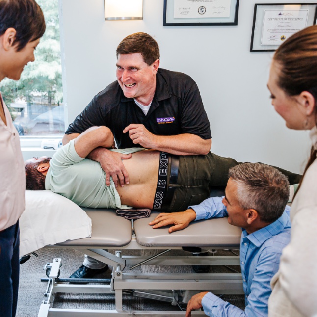 A group of practitioners gather around Bret and a patient lying down on a chiropractic drop table